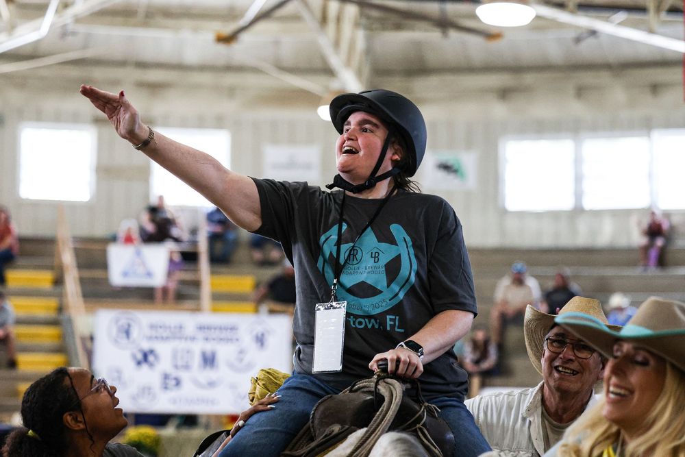 Young rider on horseback points excitedly during an indoor equestrian event, surrounded by smiling spectators.