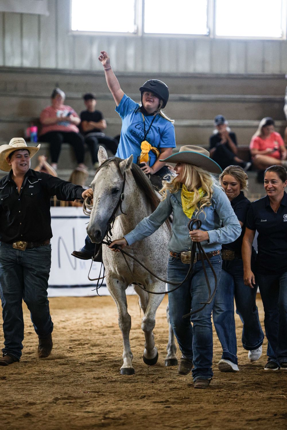 Young rider raises fist in triumph on gray horse at indoor equestrian event, assisted by handlers in cowboy hats.