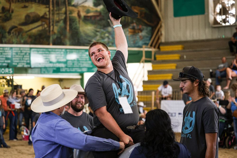 Excited man raises hat in celebration while being lifted by crowd at an indoor rodeo arena event.