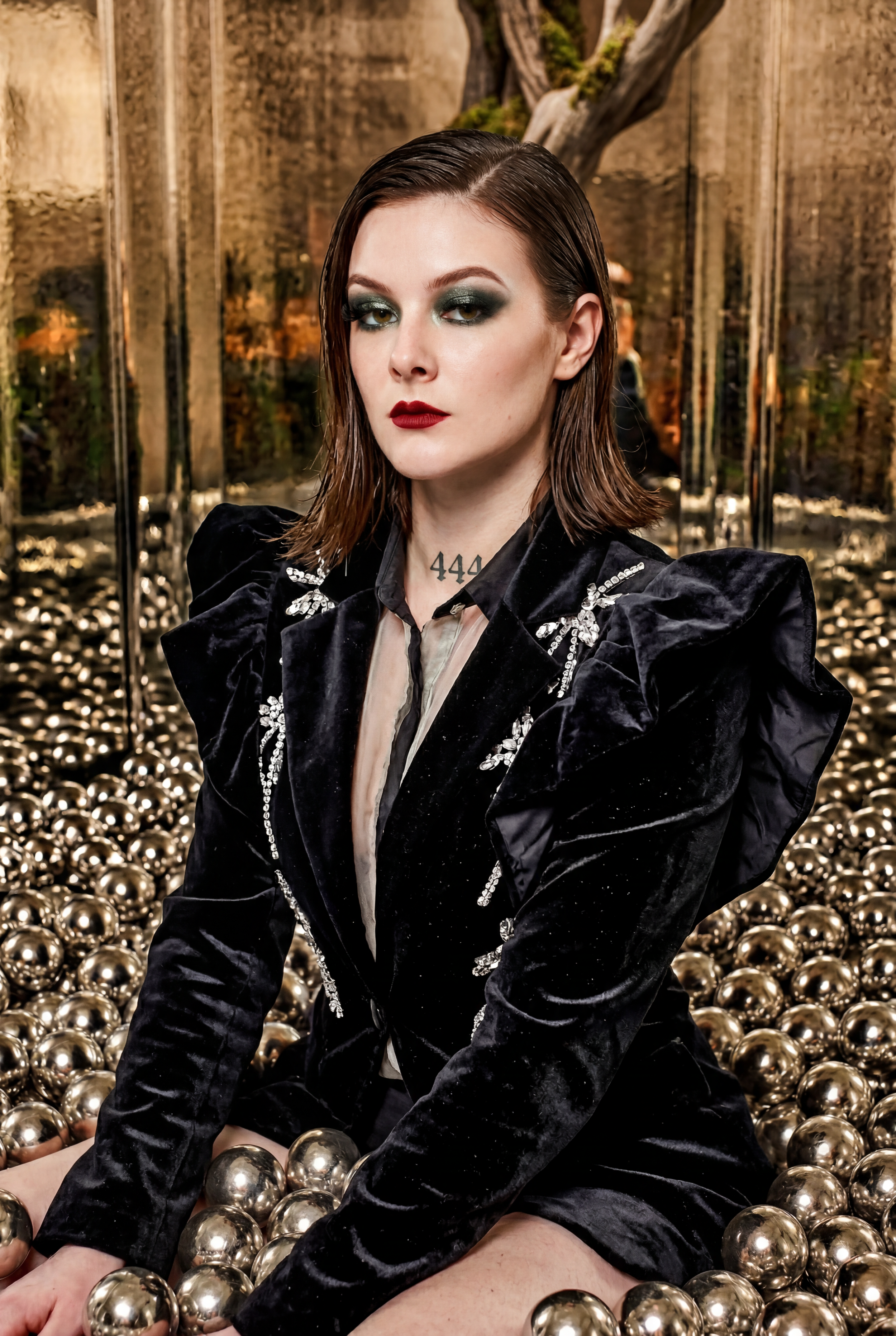 Woman in black velvet blazer with silver embroidery sits among gold metallic spheres with dramatic makeup.
