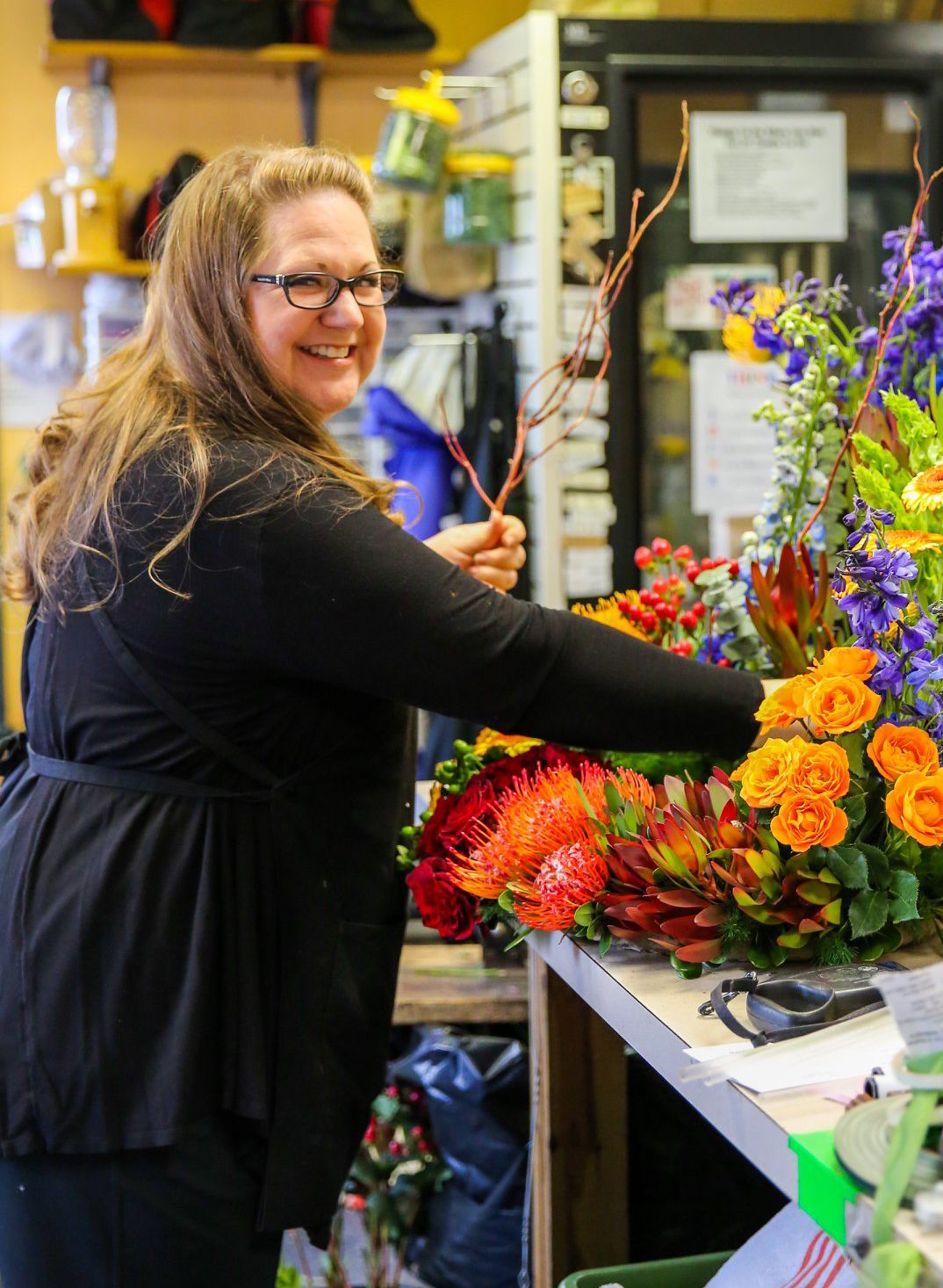 Smiling florist in black arranging colorful orange roses and purple flowers in a bright flower shop.