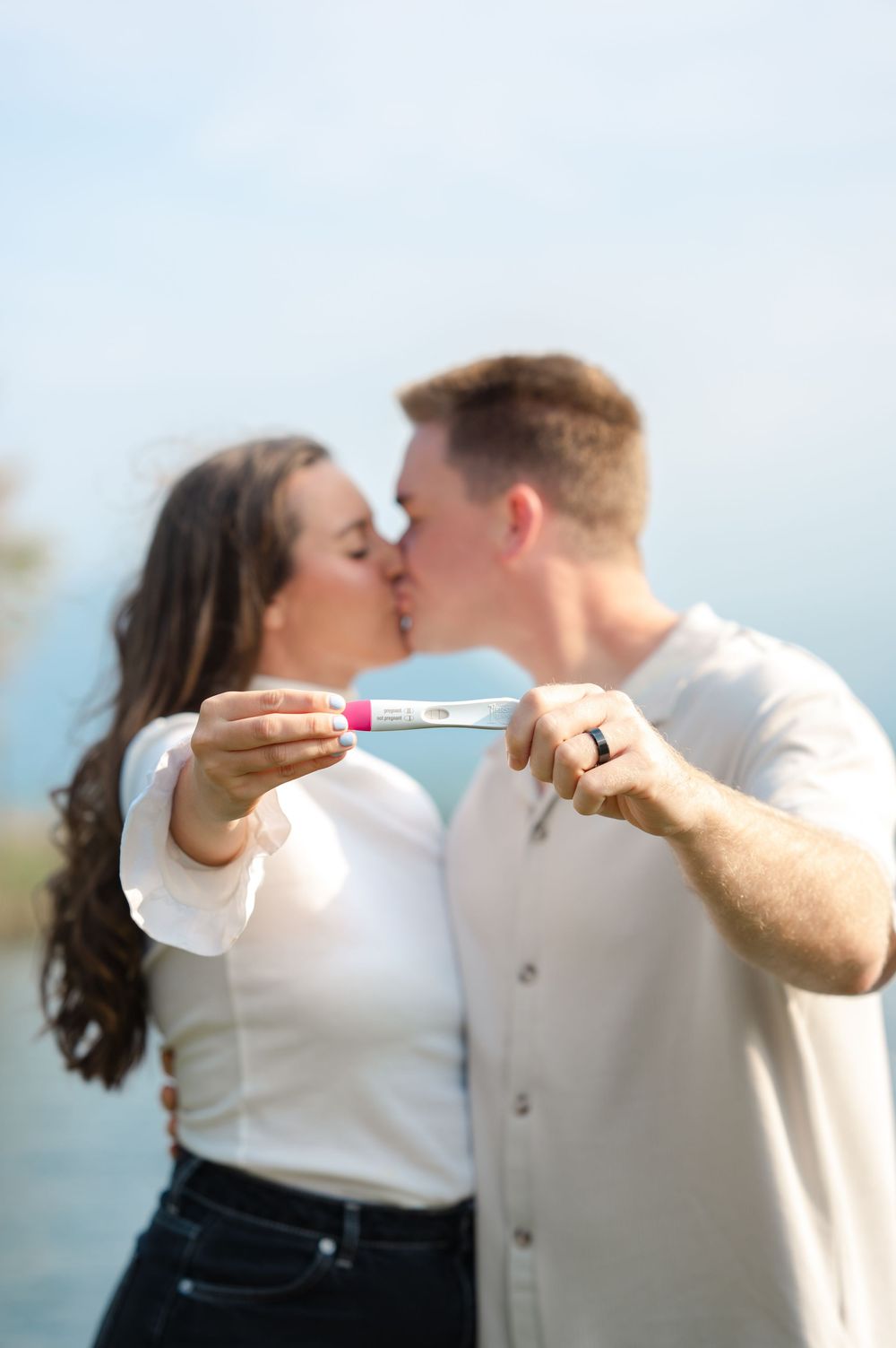 Couple kissing while holding a positive pregnancy test in front of a scenic lakeside backdrop.
