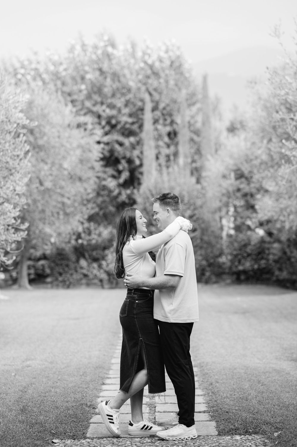 Black and white photo of a couple embracing and gazing at each other on a tree-lined path.