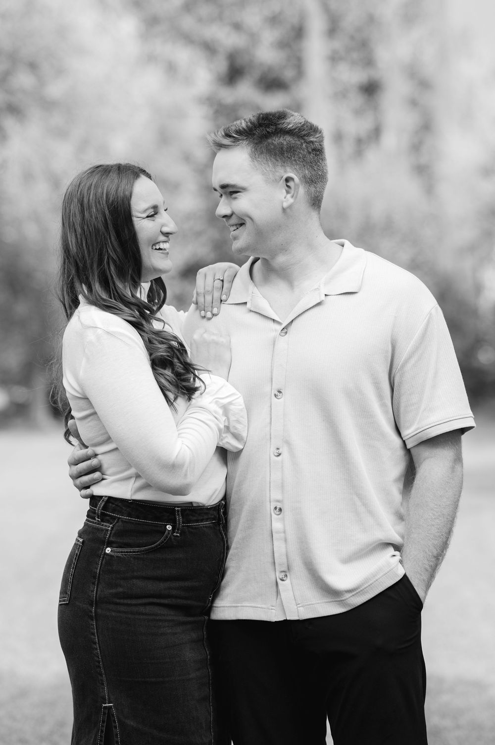 Black and white portrait of a smiling couple embracing and looking at each other in an outdoor setting.