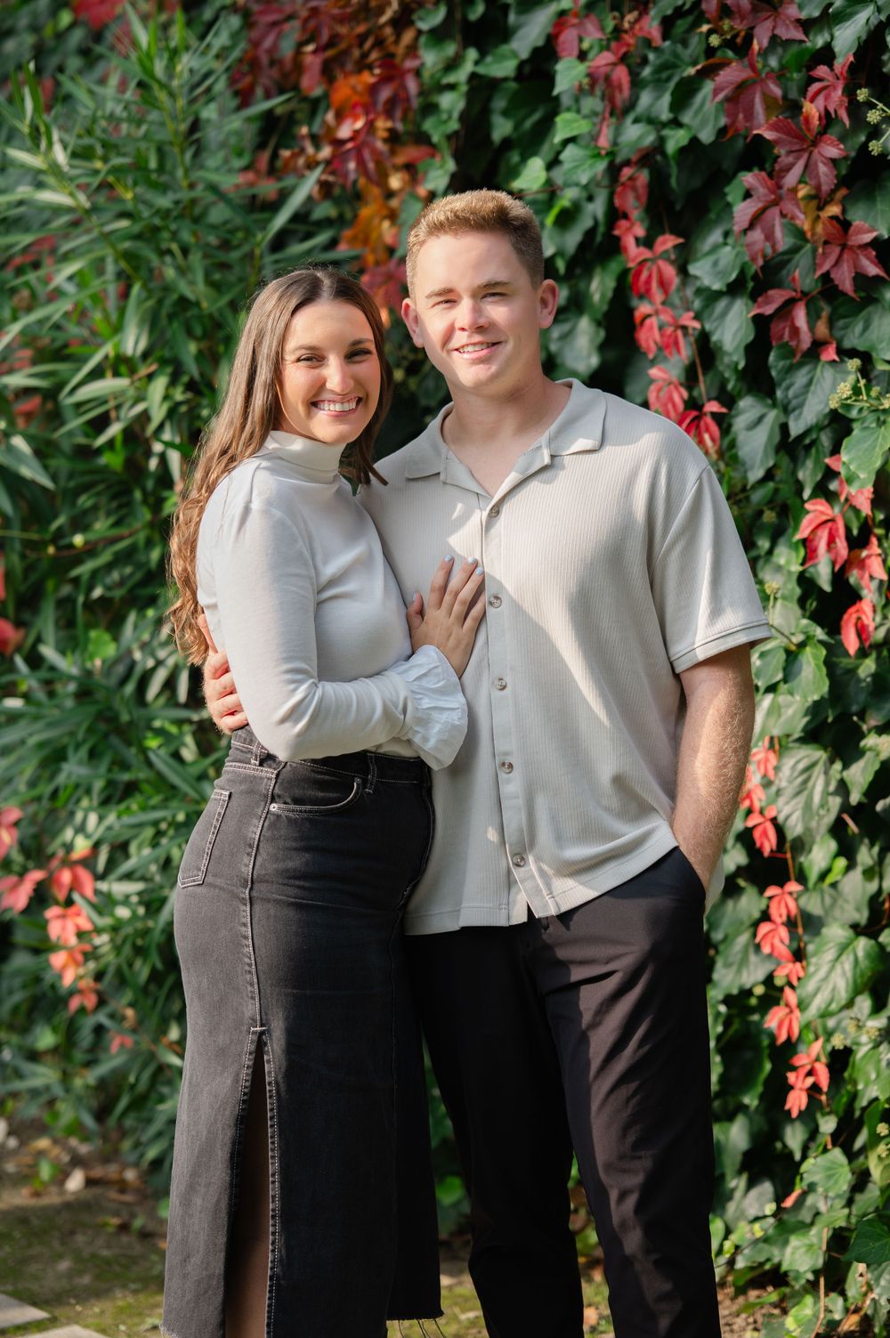A smiling couple poses together in front of a vibrant red and green ivy wall.