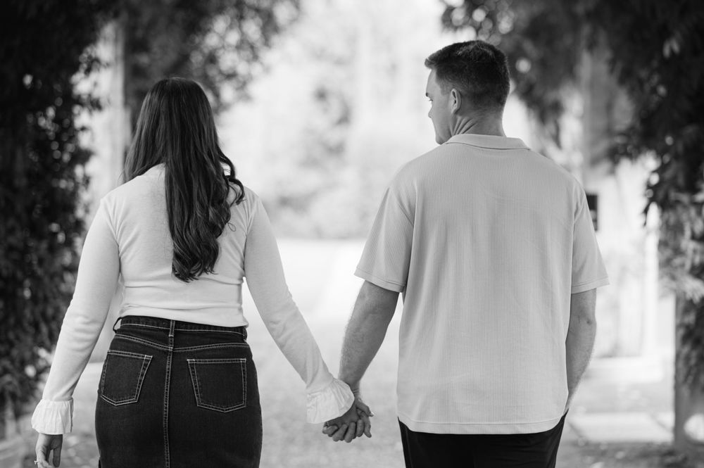 Black and white photo of a couple holding hands walking along a tree-lined garden path.
