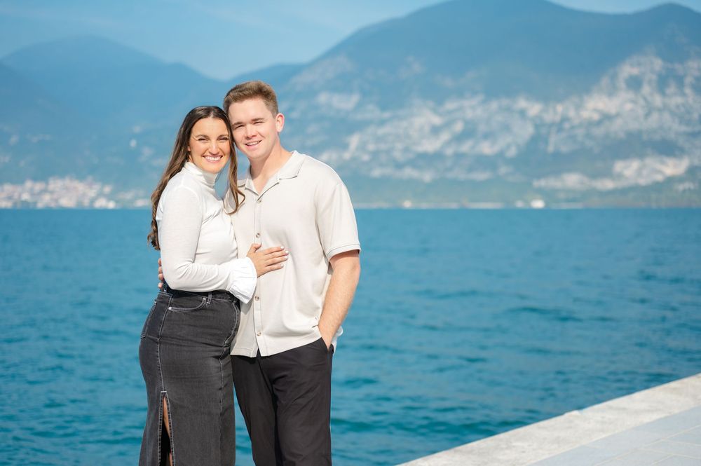 Smiling couple posing together on a waterfront pier with blue lake and mountain backdrop.