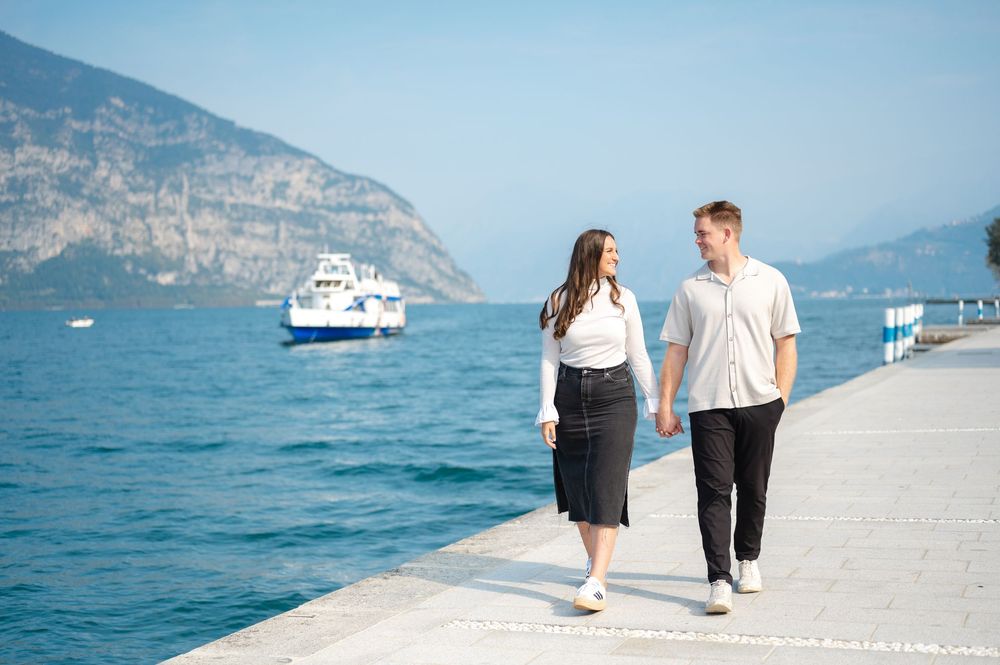 Happy couple walking hand in hand along a sunny lakeside pier with mountains and a ferry boat.