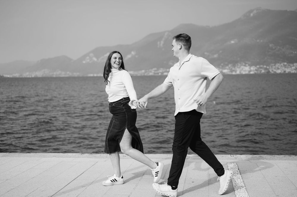 Black and white photo of a happy couple holding hands and walking along a waterfront promenade with mountains in the background.