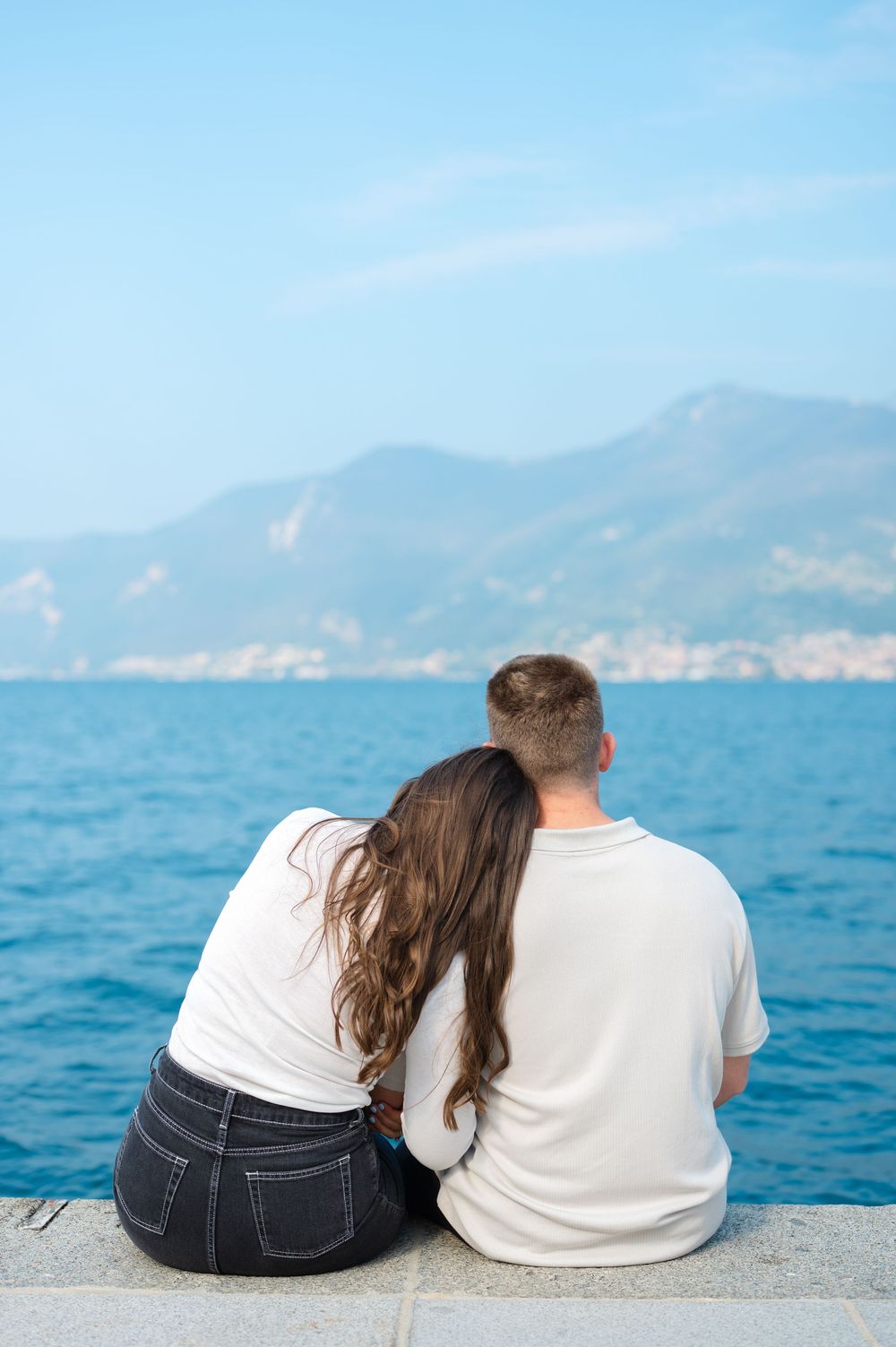 A couple sits side by side on a waterfront ledge gazing at a scenic mountain lake view.