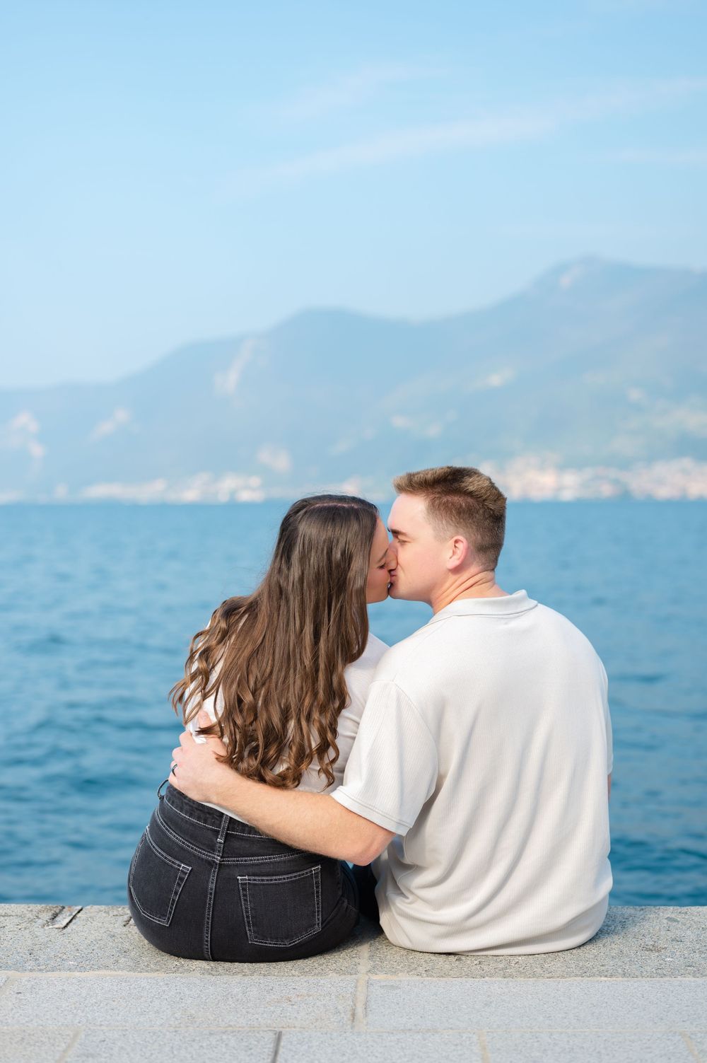 Couple sharing a kiss while sitting on a waterfront wall with mountain views.