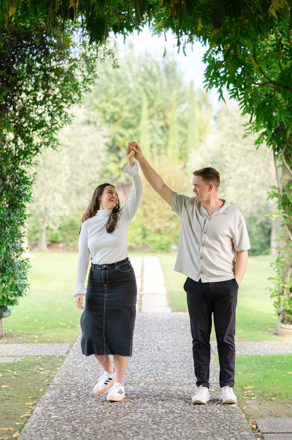 A couple dances joyfully under a green leafy archway on a garden path.