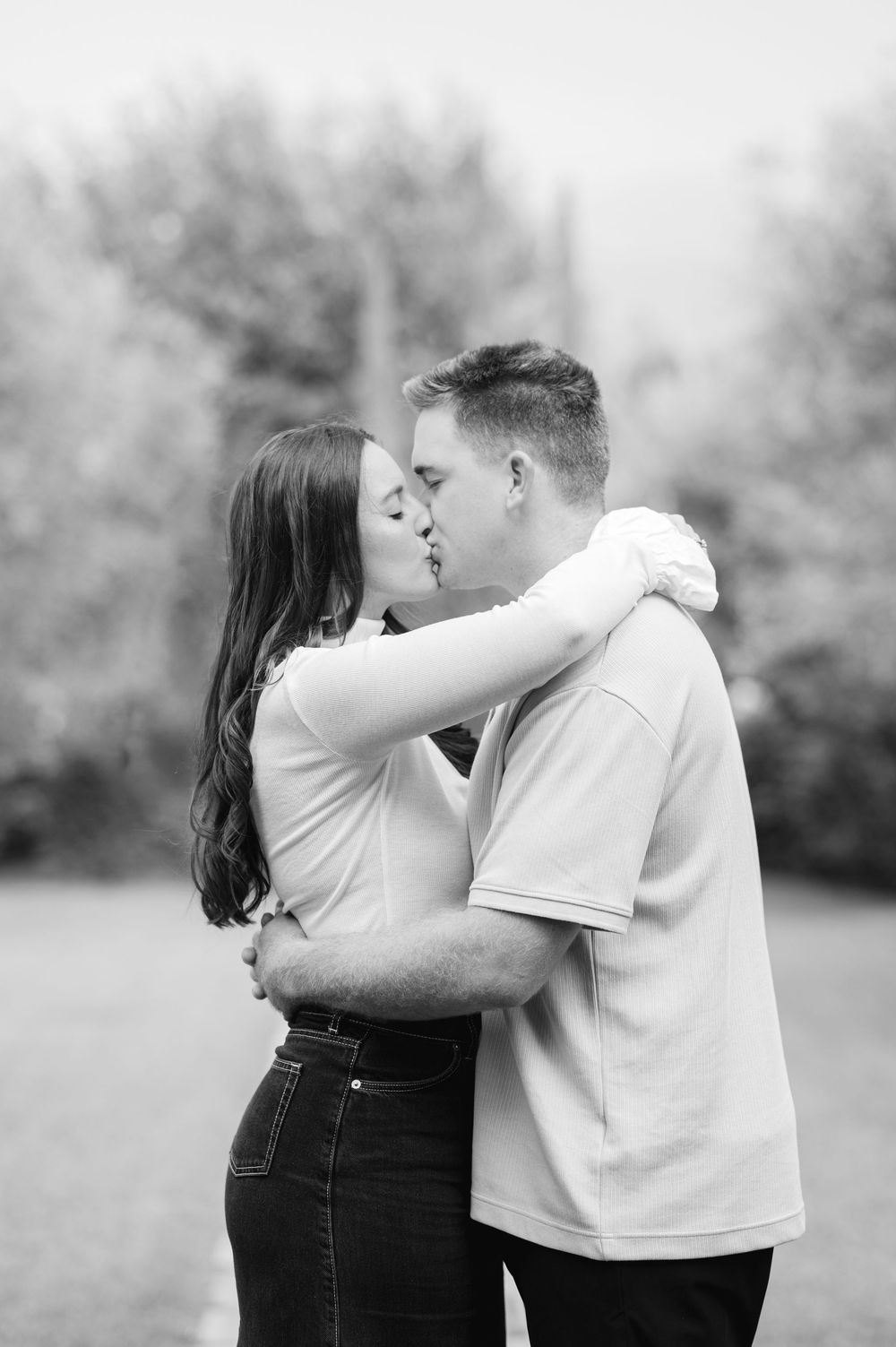 Black and white photo of a couple sharing a romantic kiss outdoors with blurred trees in the background.