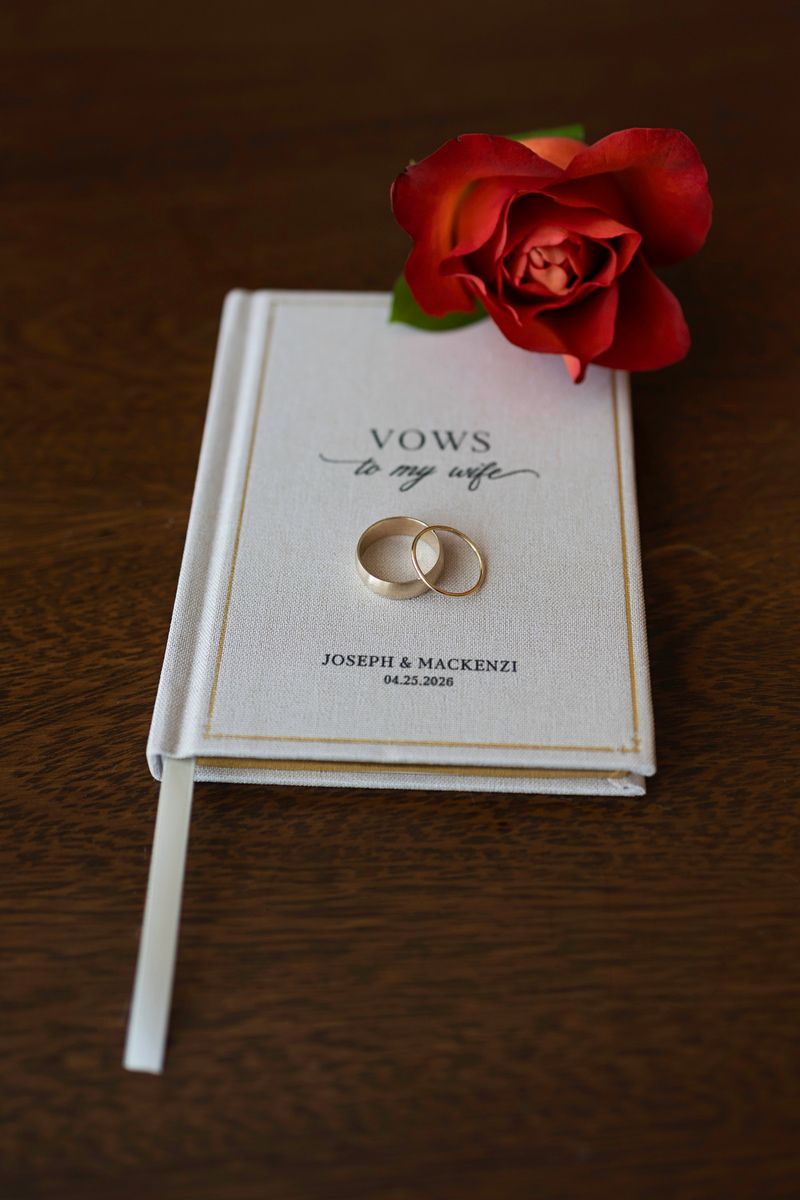 Gold wedding rings rest on a personalized vows book with a red rose, reading Joseph & Mackenzi 04.25.2026.