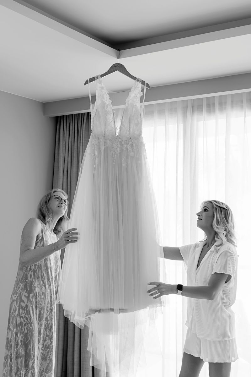 Two women admire a hanging wedding dress in a bright room, captured in elegant black and white photography.