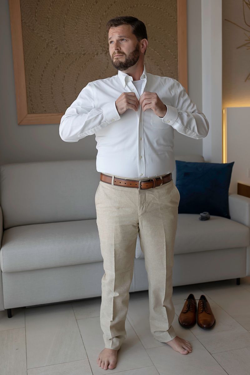 Man in white shirt and beige pants buttoning his collar while standing barefoot in a modern living room.