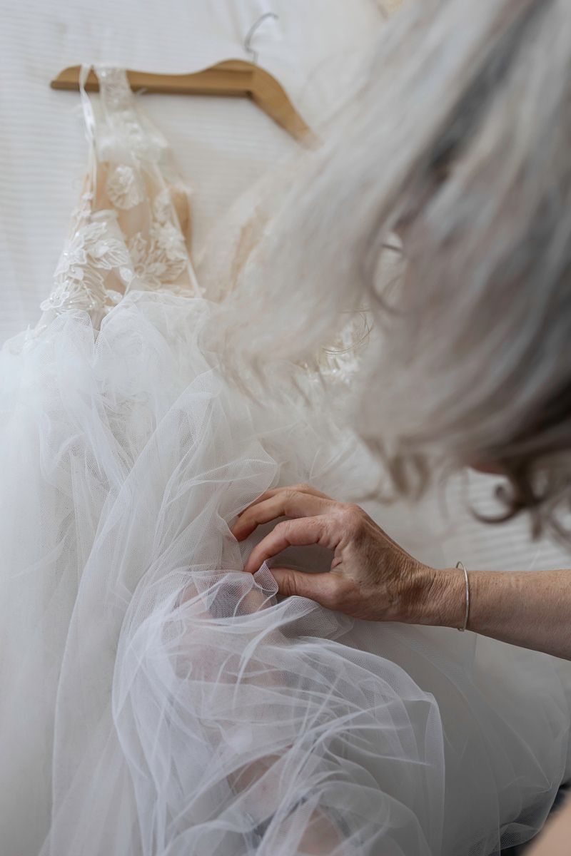 Woman with gray hair touching delicate white tulle wedding dress hanging on wooden hanger in bridal boutique.