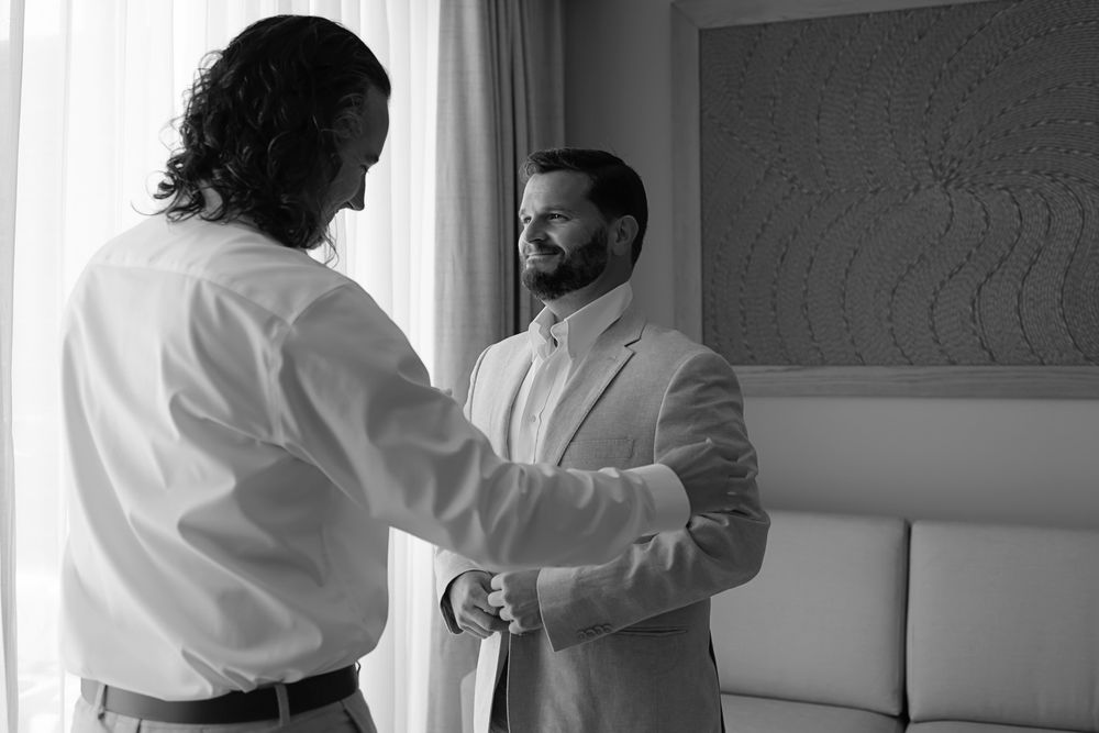 Black and white photo of two men in formal attire helping each other prepare for a wedding ceremony indoors.