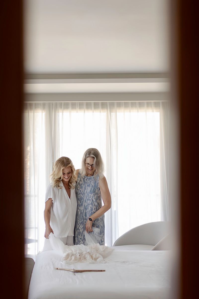 Two blonde women smile while looking at a wedding dress laid out on a bed in a bright hotel room.