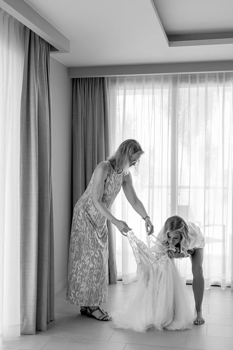 Black and white photo of a woman helping a bride into her wedding dress near a window.