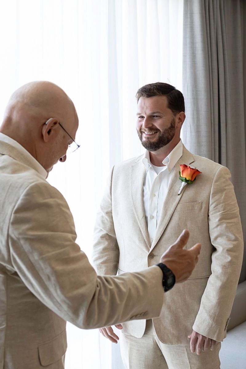Smiling groom in beige linen suit with orange rose boutonniere chatting with bald man in matching suit before wedding.