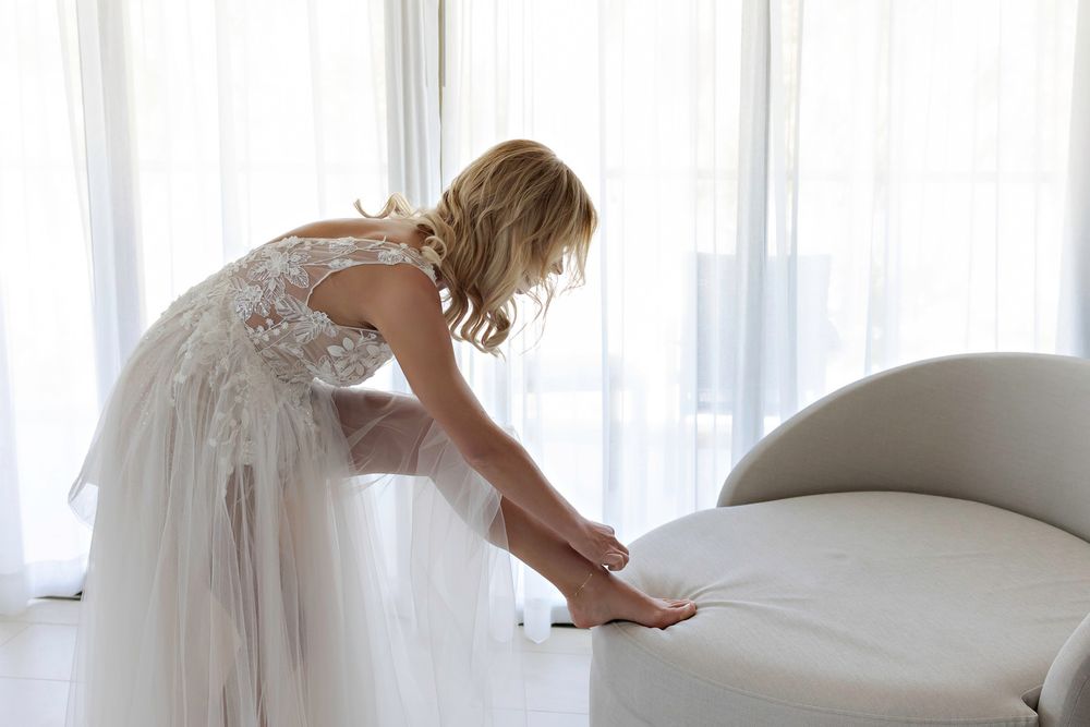 Bride in white lace wedding dress adjusting her heels on a chaise lounge by sheer curtained windows.