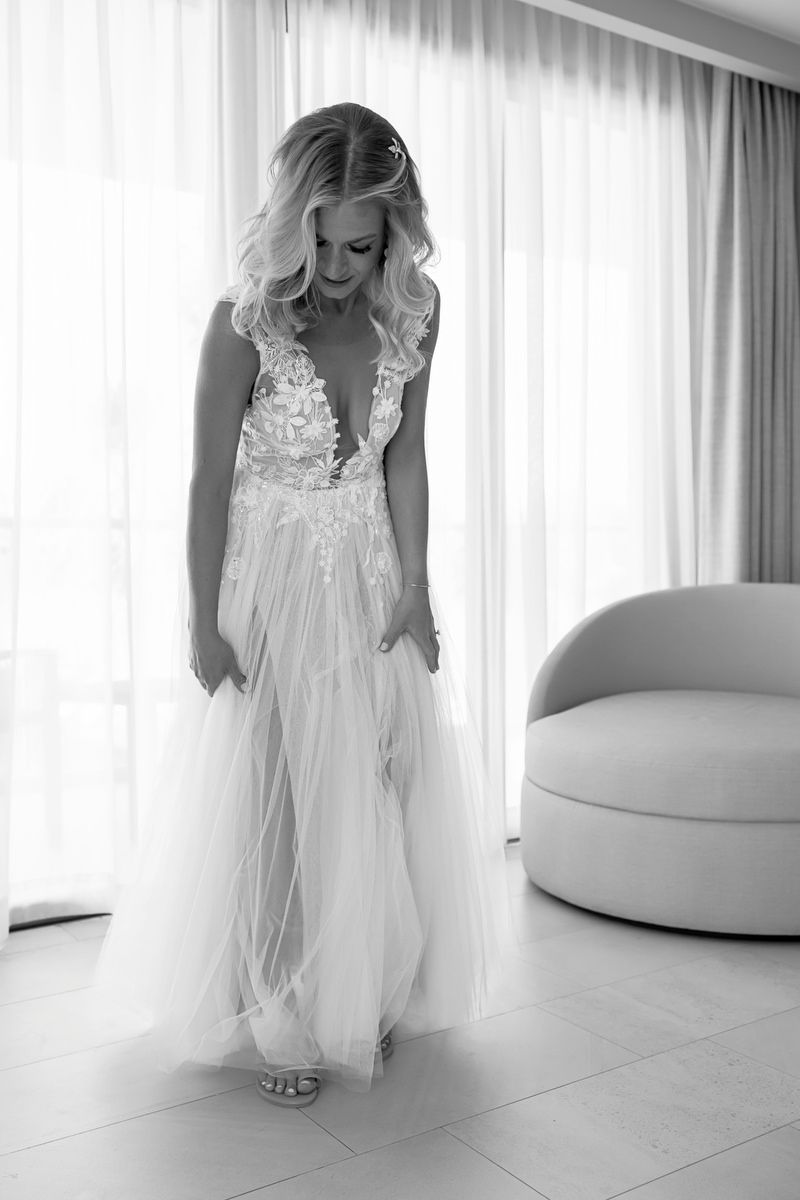 Black and white photo of a bride in a stunning deep V-neck lace tulle wedding gown, posing elegantly by sheer curtains.