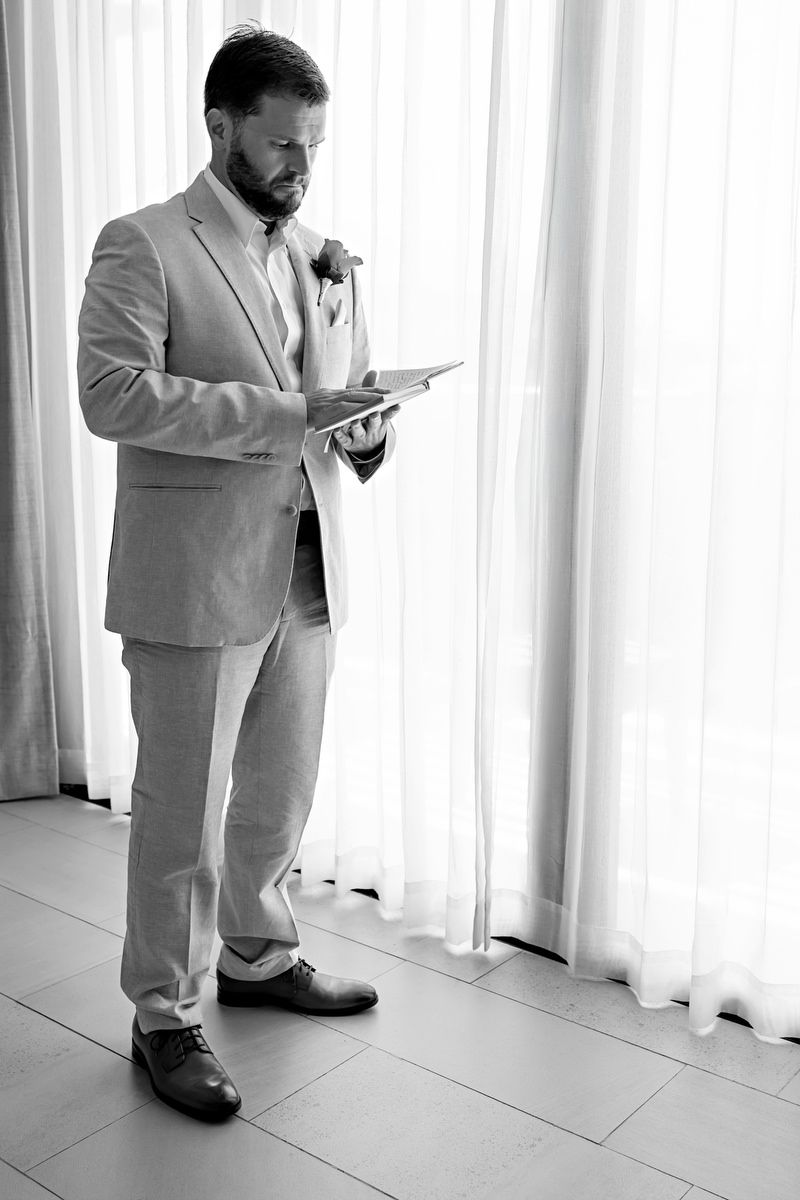 Groom in beige linen suit reading wedding vows by sheer curtained window in black and white photo.