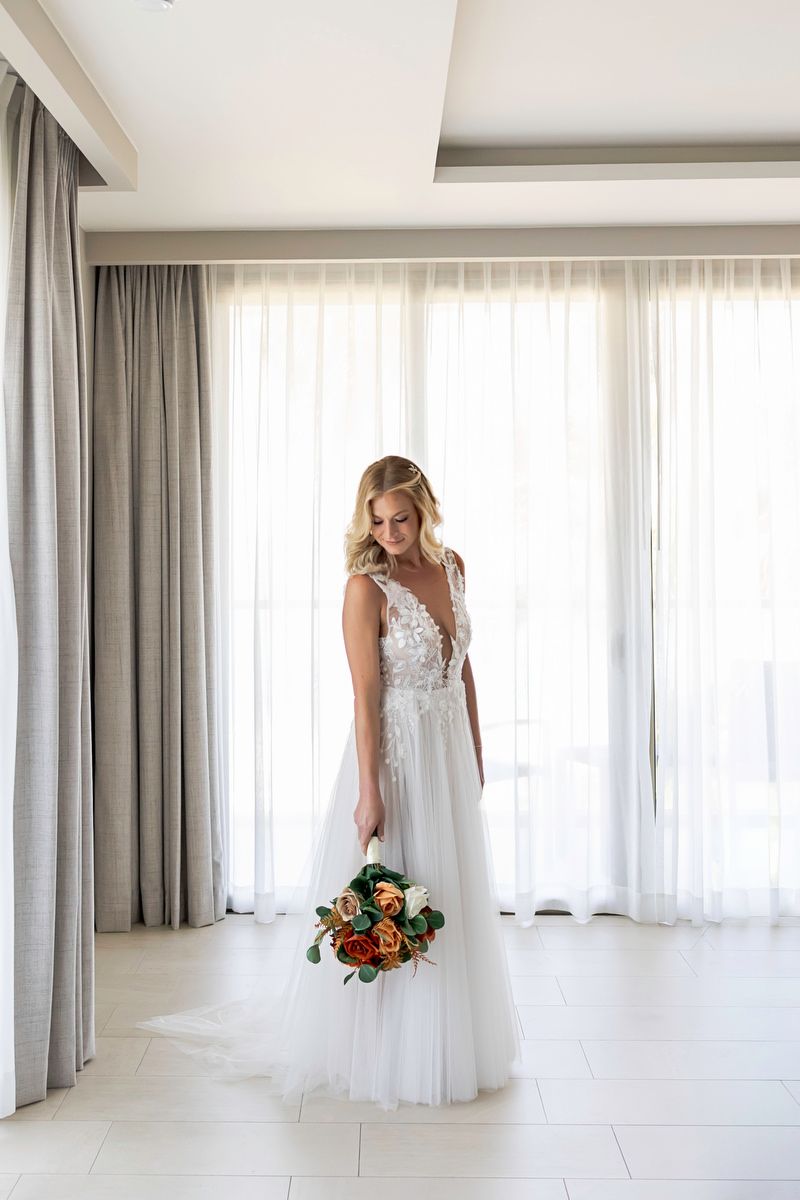 Bride in white lace tulle gown holds colorful bouquet of red and orange roses, standing by sheer curtains in bright room.