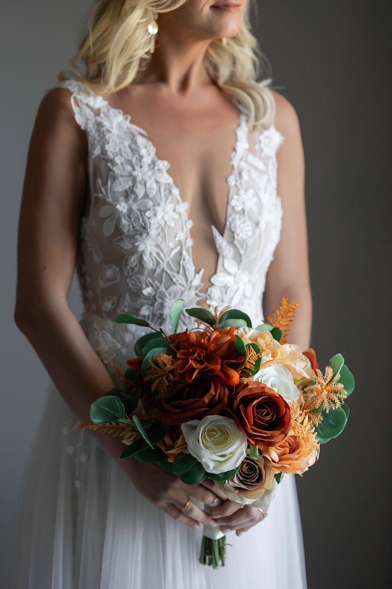 Bride in white lace plunging neckline gown holding autumn bouquet with burnt orange, cream roses and greenery.