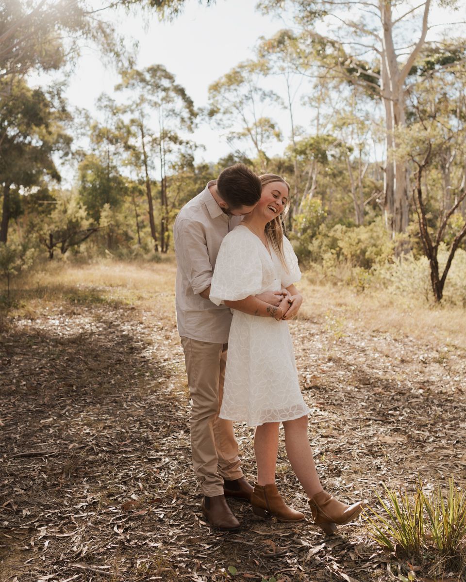 Couple embracing and laughing in a sunlit Australian bush setting, woman in white dress and brown boots.