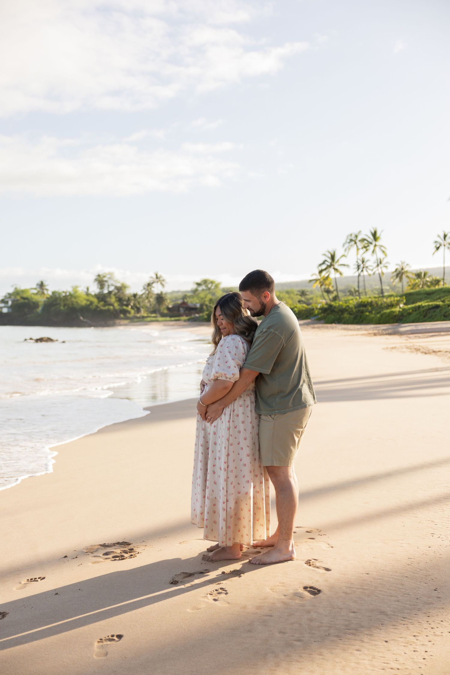Couple embracing on a sandy Hawaiian beach at sunset with lush green trees and volcanic rocks in the background.