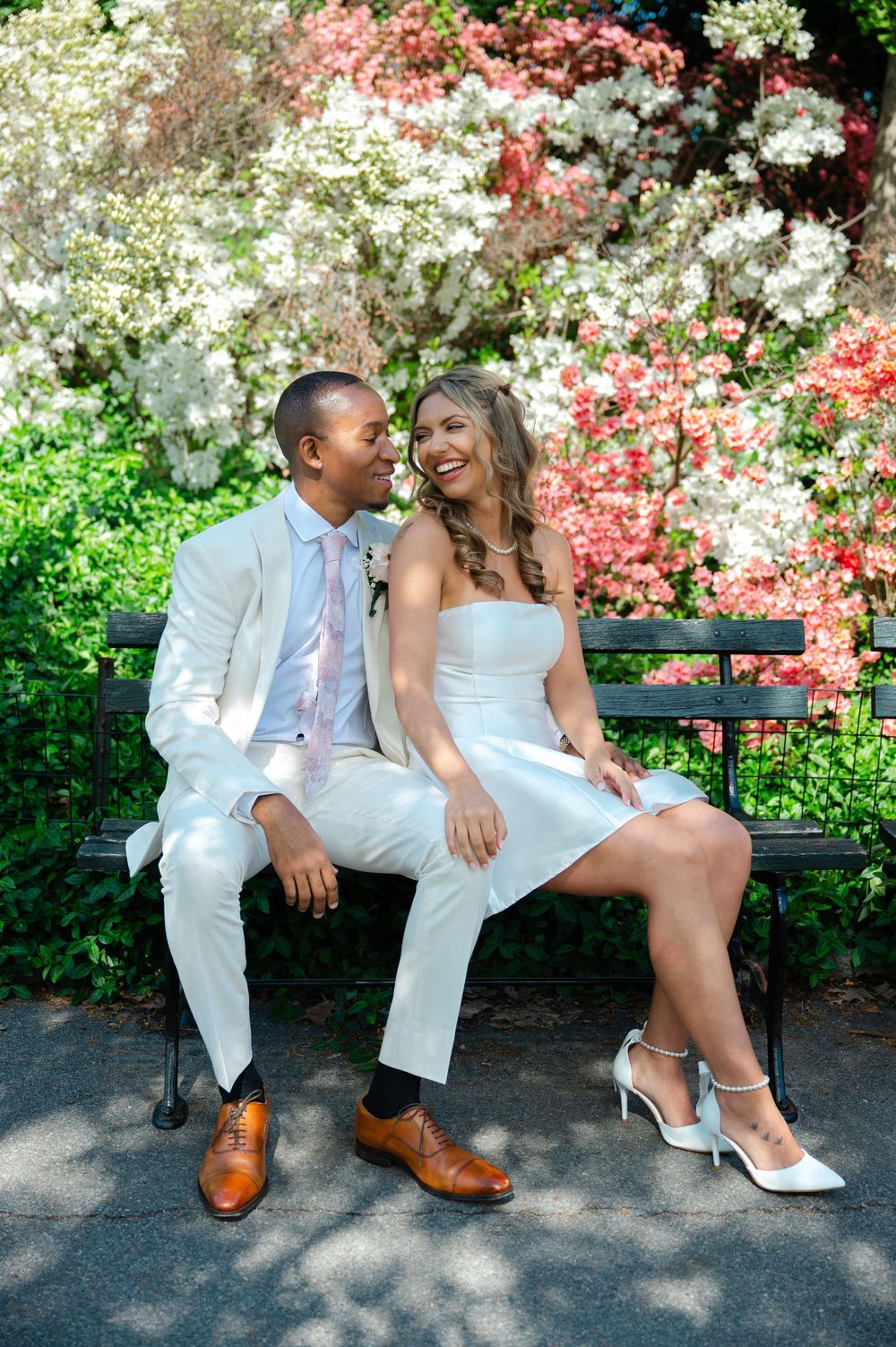 Bride and groom in white wedding attire sitting on park bench surrounded by colorful spring flowers.