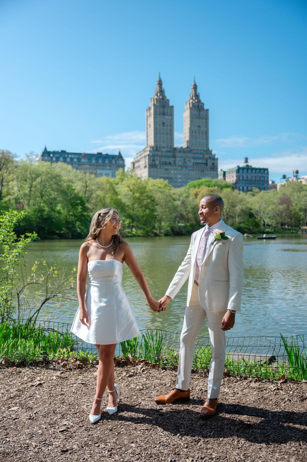 Newlywed couple holds hands by Central Park lake with iconic San Remo building in background on sunny day.