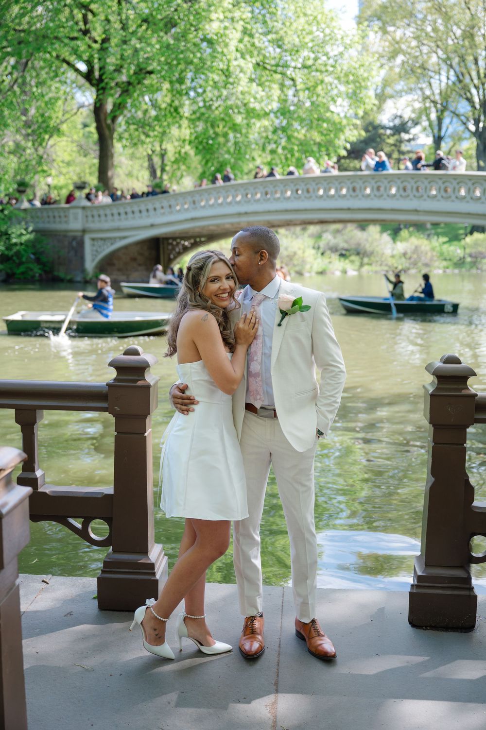 Couple in wedding attire poses by Bow Bridge in Central Park, NYC, with rowboats on the lake behind them.