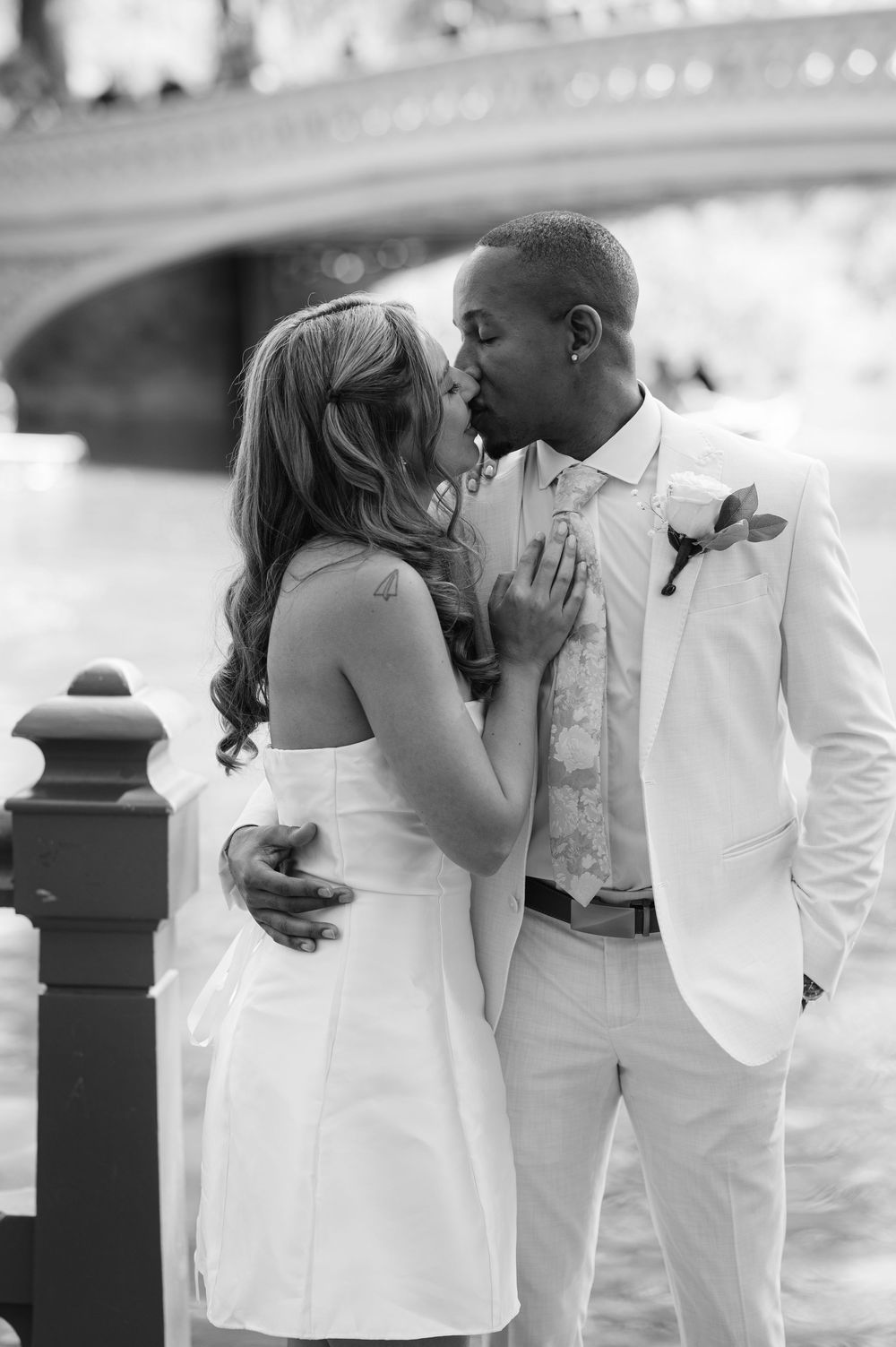 Black and white photo of an interracial couple sharing a kiss in wedding attire near a bridge.