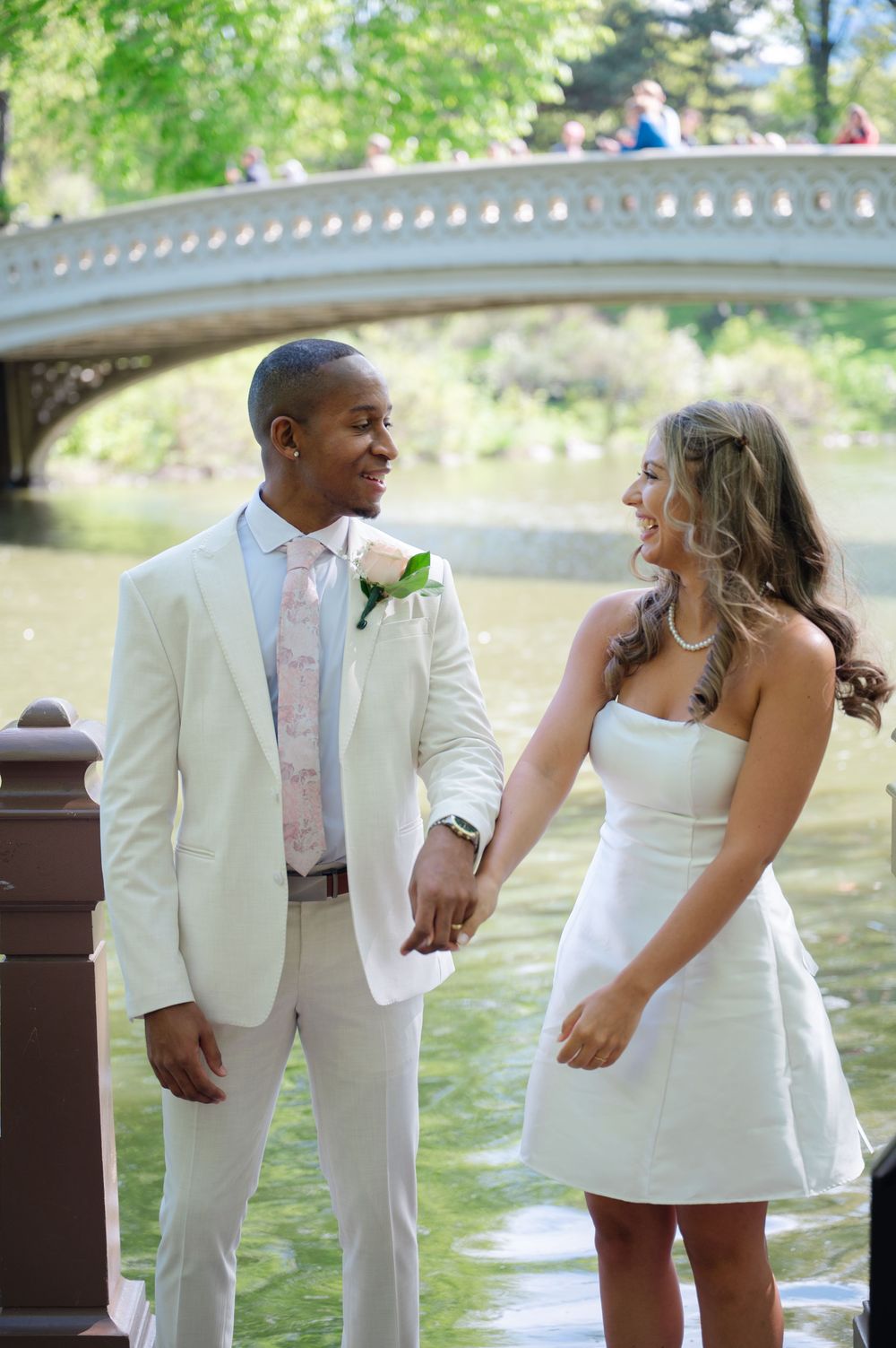 Couple in white wedding outfits holding hands near Bow Bridge in Central Park, New York City.