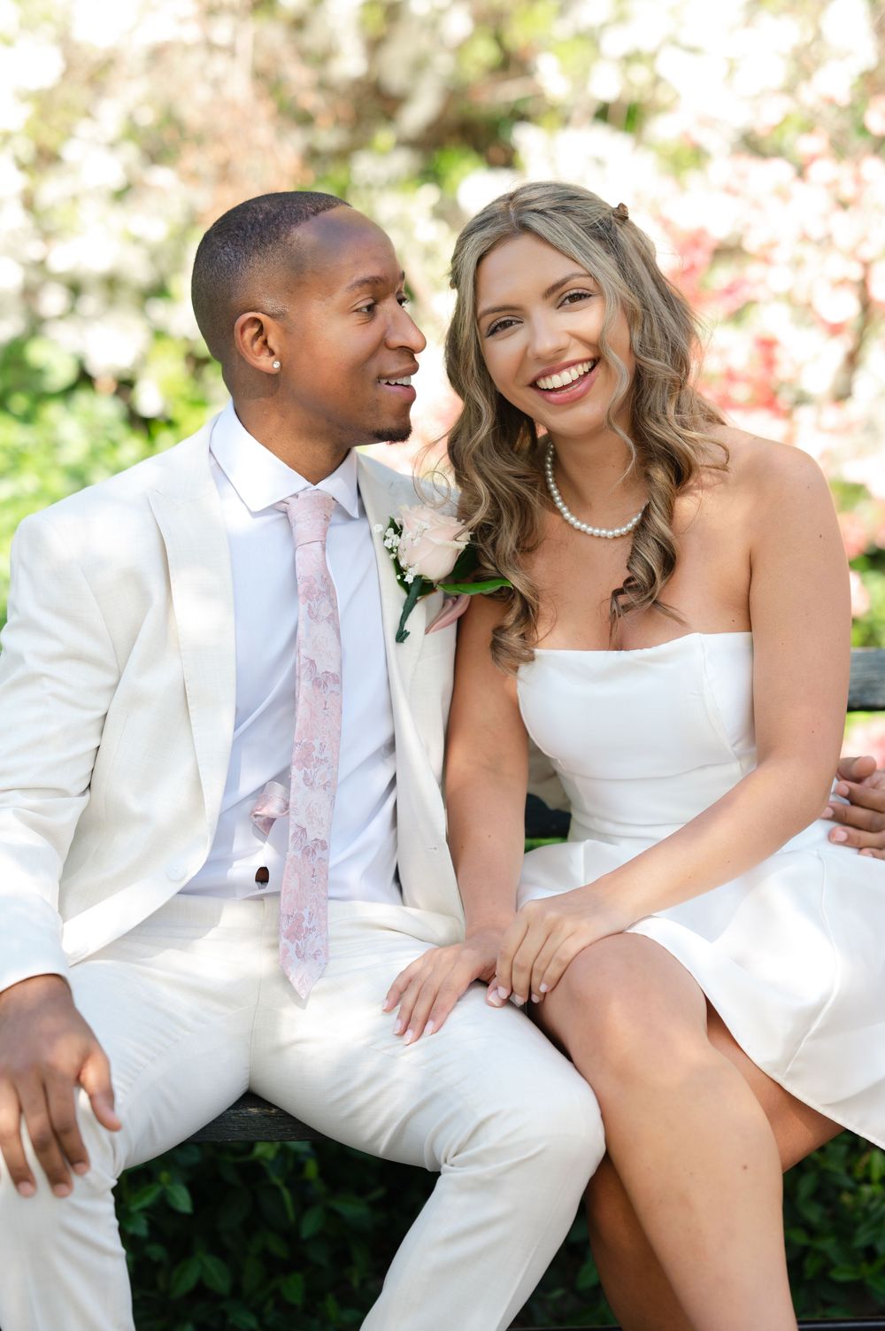 Smiling newlywed couple in white wedding attire sitting on a bench surrounded by blooming flowers.