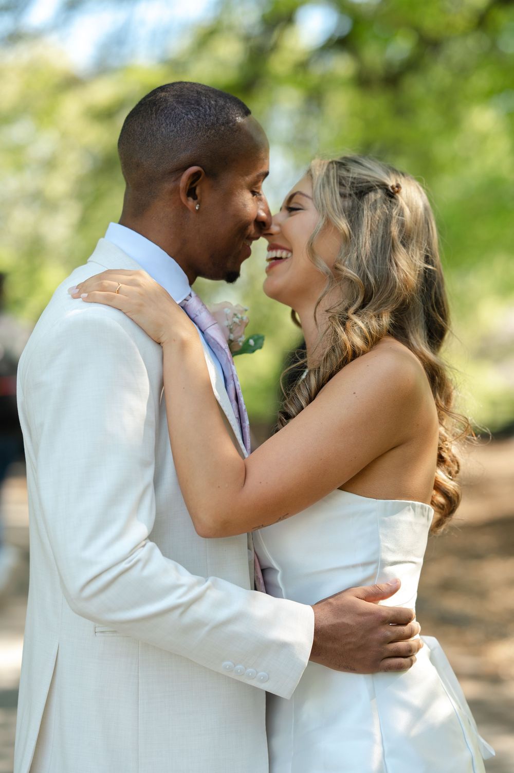 Interracial couple in white wedding attire sharing a romantic moment outdoors, smiling and embracing in a sunlit park.