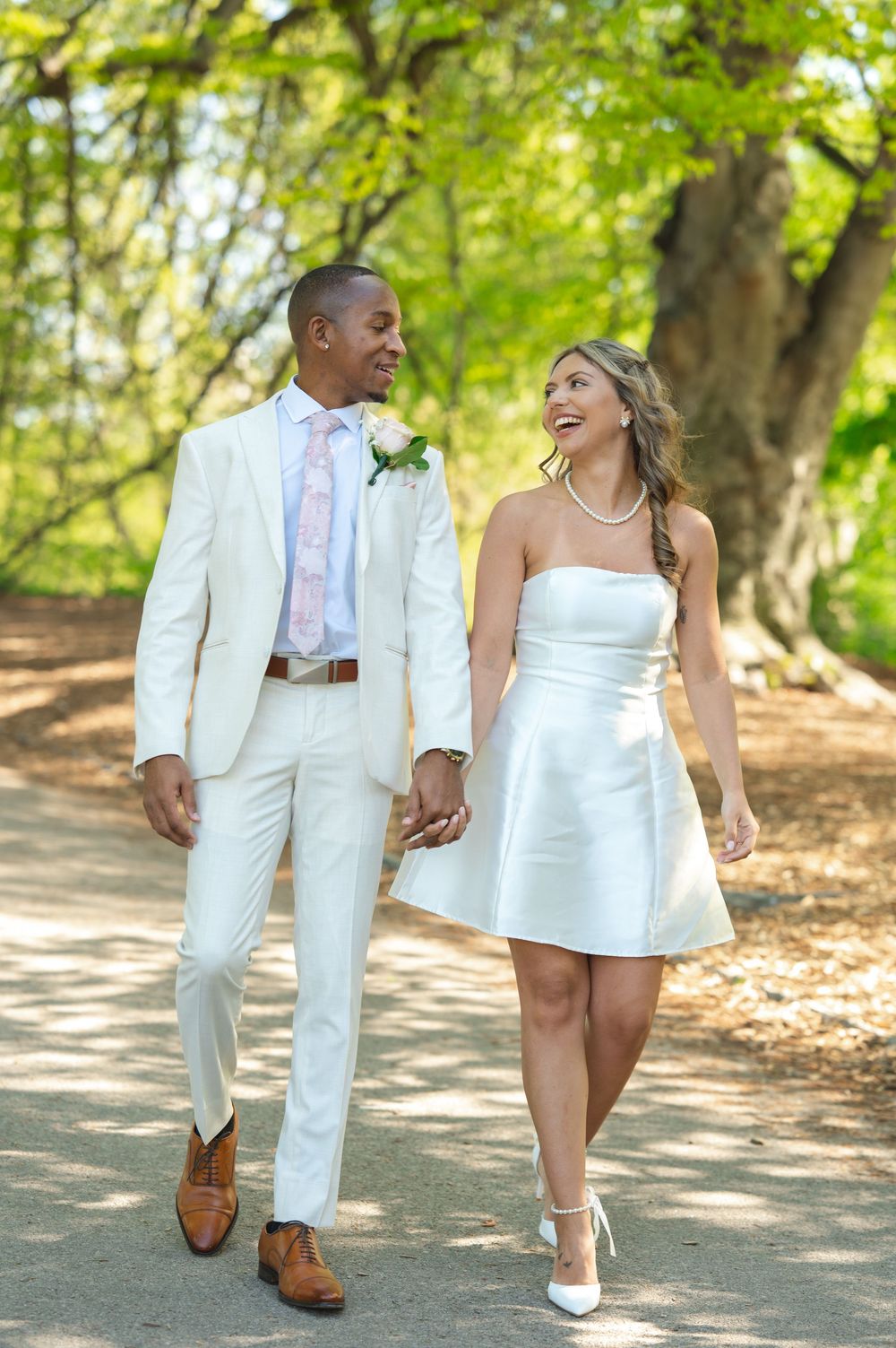 Happy interracial newlywed couple walking hand in hand on a sunlit park path in white wedding attire.