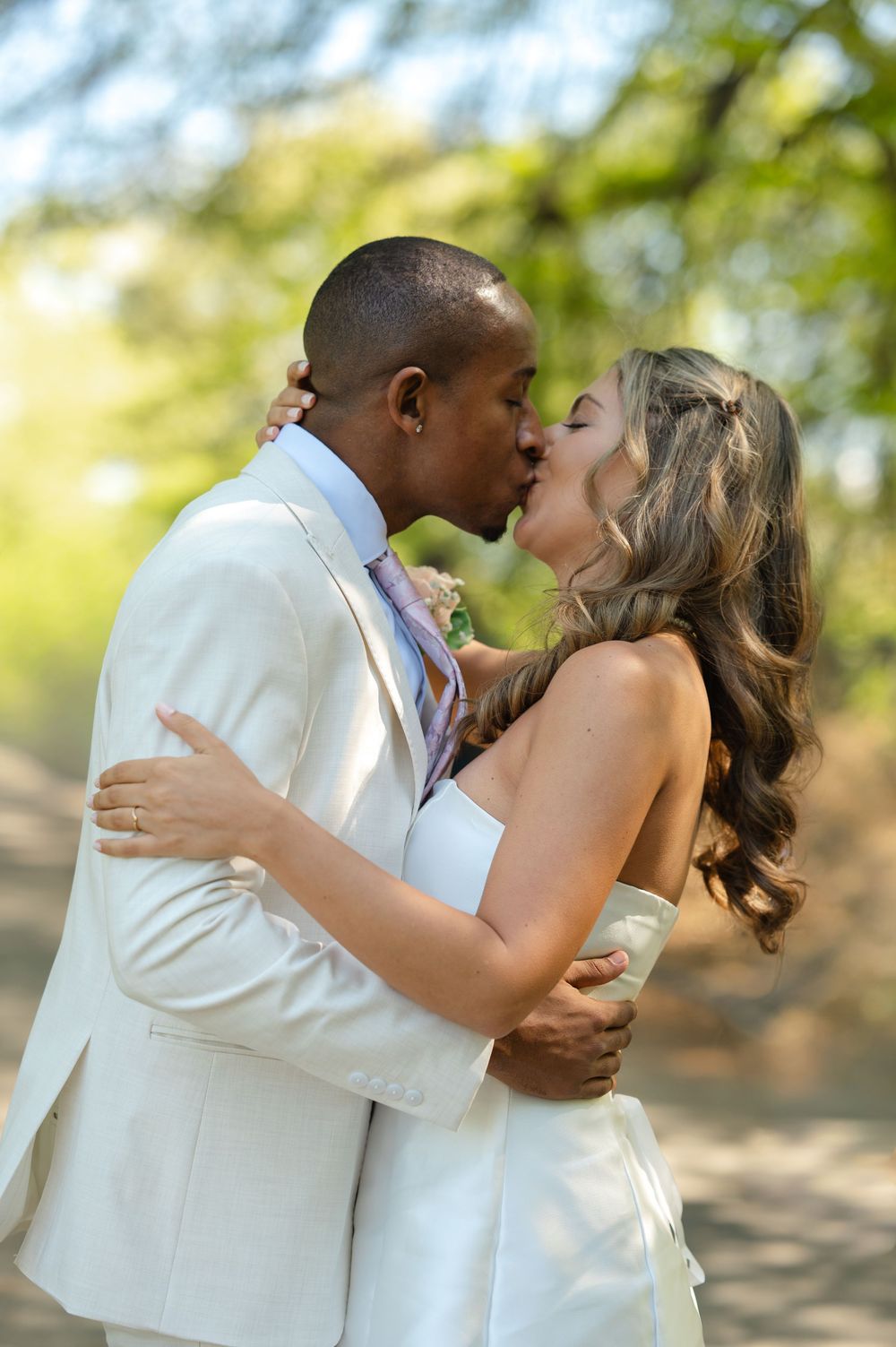 Interracial bride and groom share first kiss outdoors in lush green park setting.