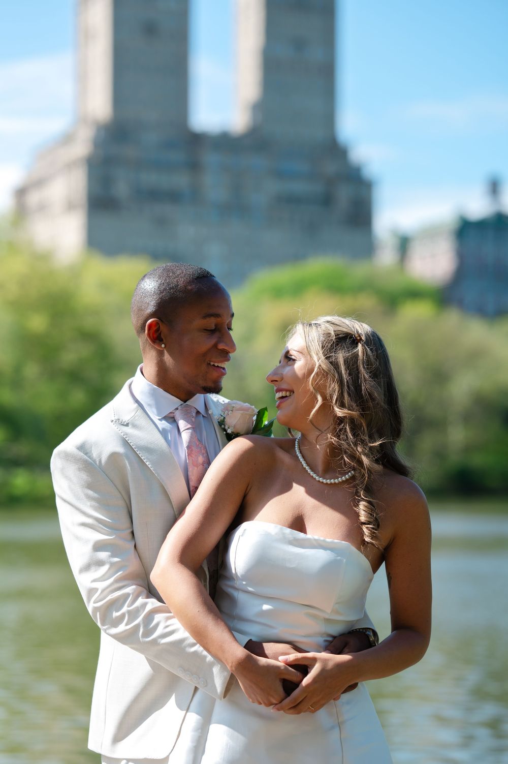 Interracial couple in wedding attire smiling at each other in Central Park with a lake and buildings in the background.