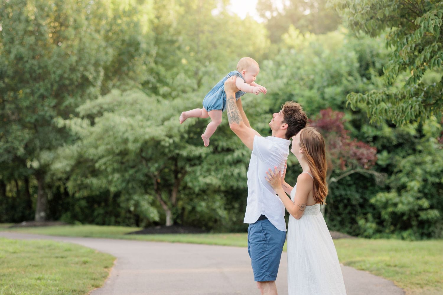 Happy family in a park, father lifting baby in the air while mother watches, surrounded by lush green trees.