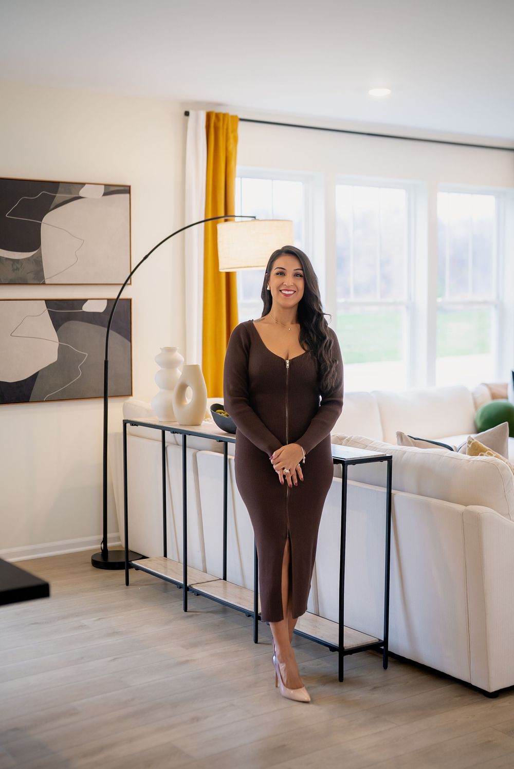 Woman in brown midi dress and heels poses confidently in modern living room with yellow curtains and arc floor lamp.