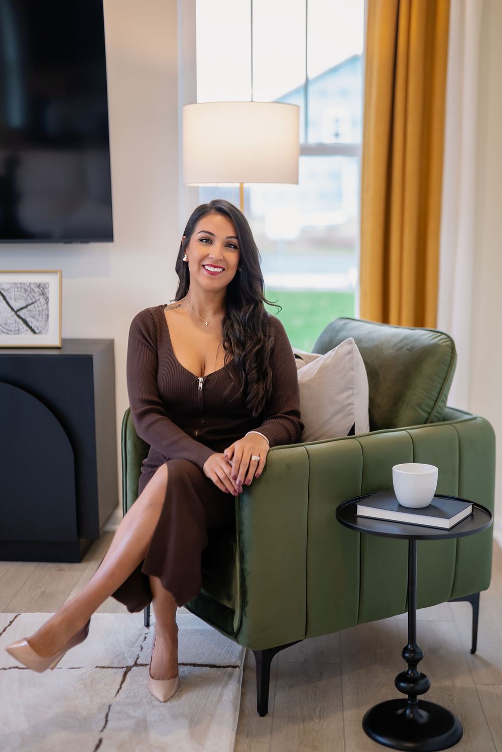 Woman in brown dress sitting elegantly in green velvet chair beside side table in modern living room.