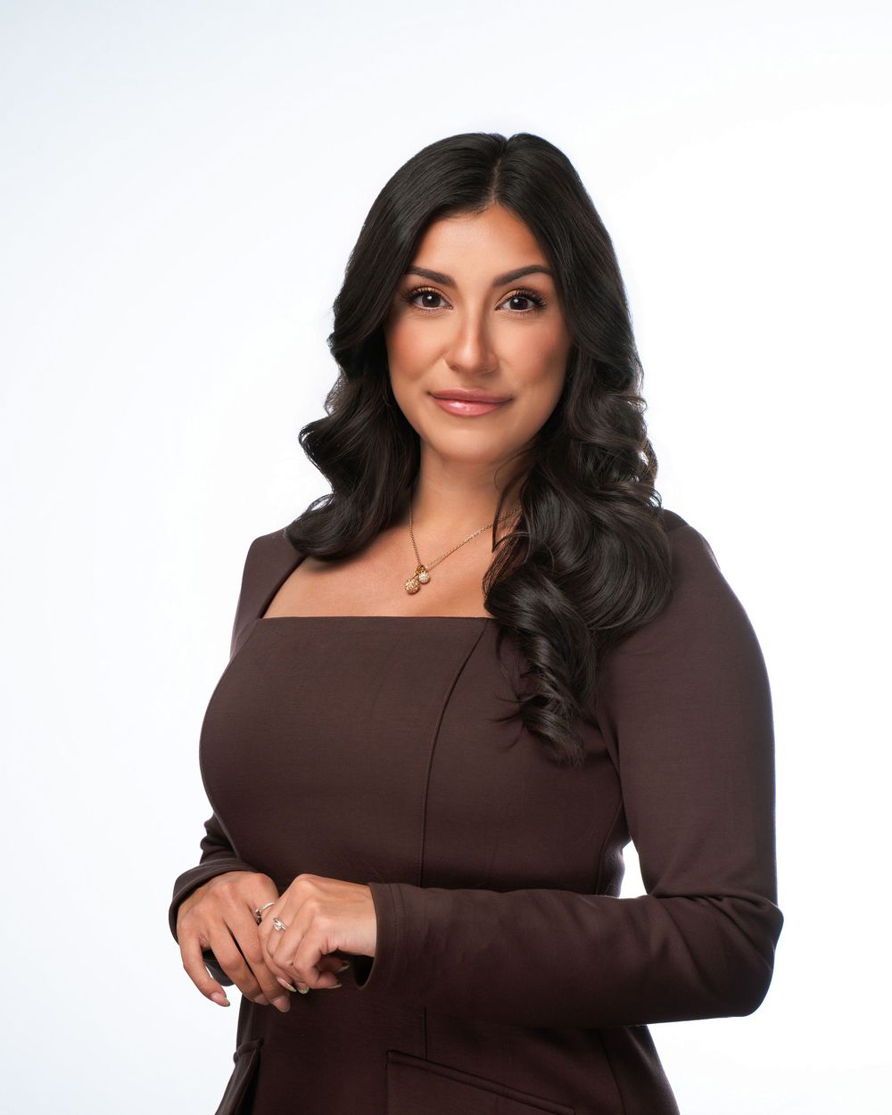 Professional woman with dark wavy hair wearing a brown fitted dress and gold necklace against white background.