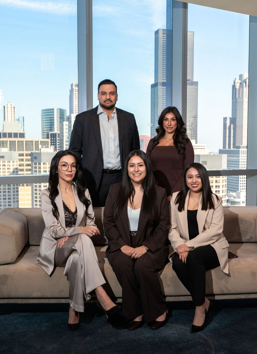 Five professional colleagues pose for a corporate team photo in a modern high-rise office with city skyline views.