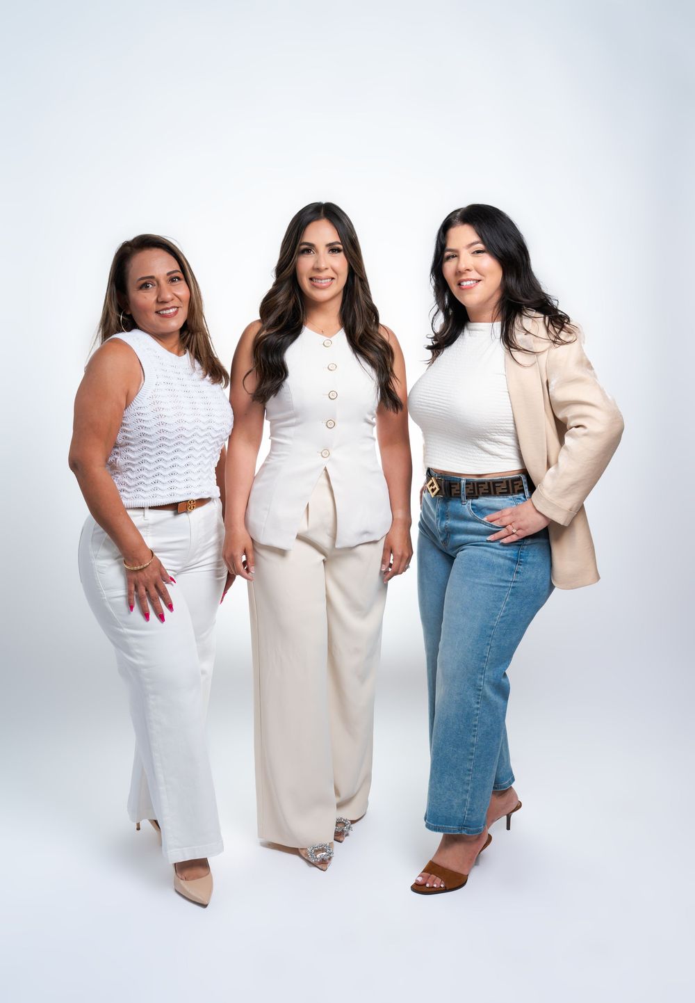 Three stylish women posing together in white and neutral outfits against a light background.