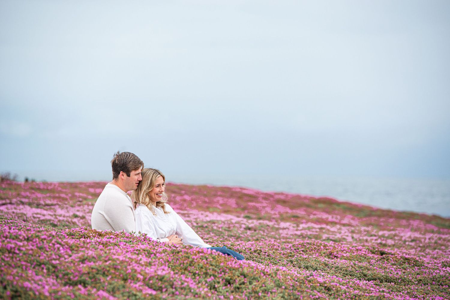 Couple sitting together in a vast field of pink wildflowers under a cloudy sky.