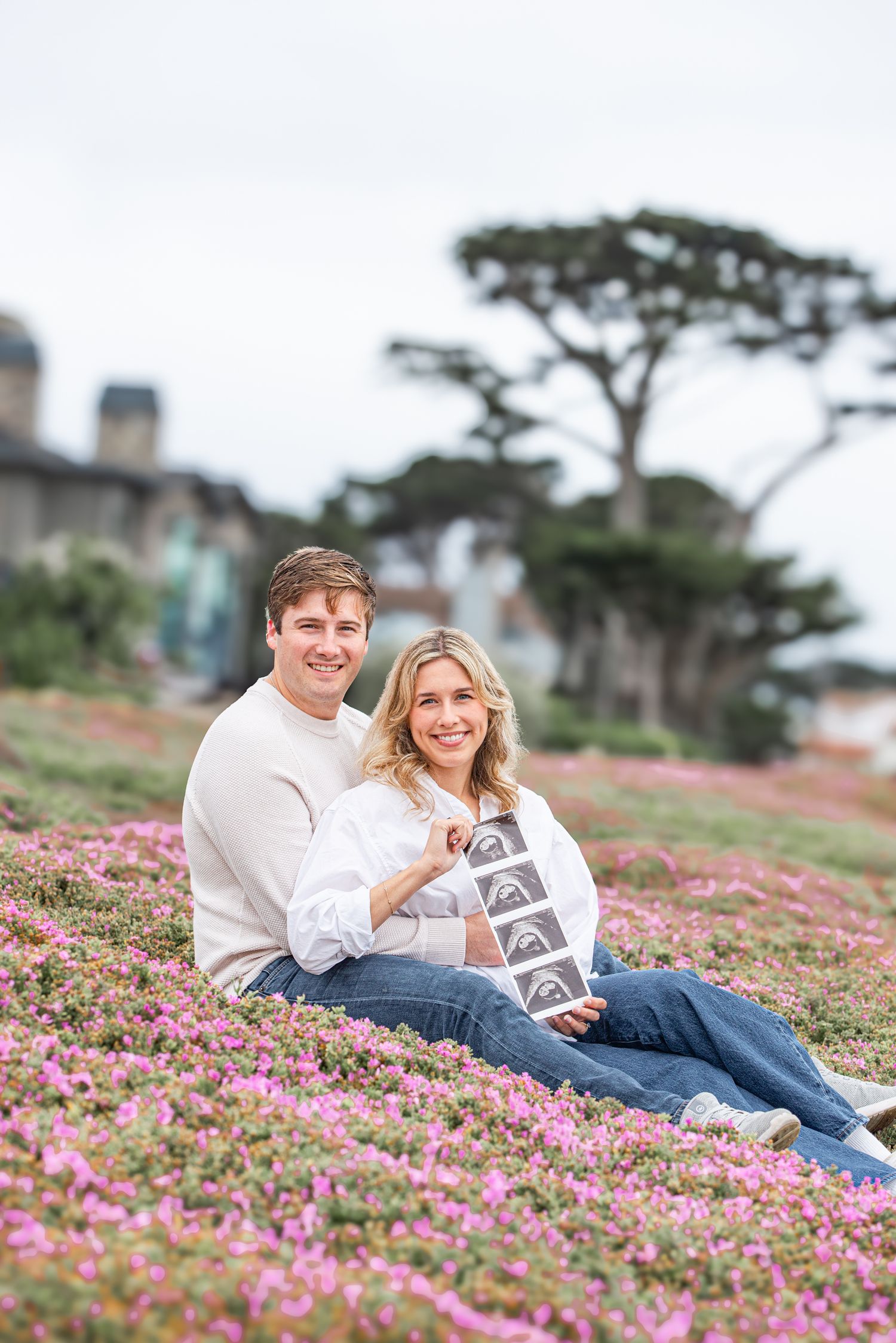 Smiling couple sitting in pink wildflowers holding ultrasound photos, announcing pregnancy outdoors near trees and building.
